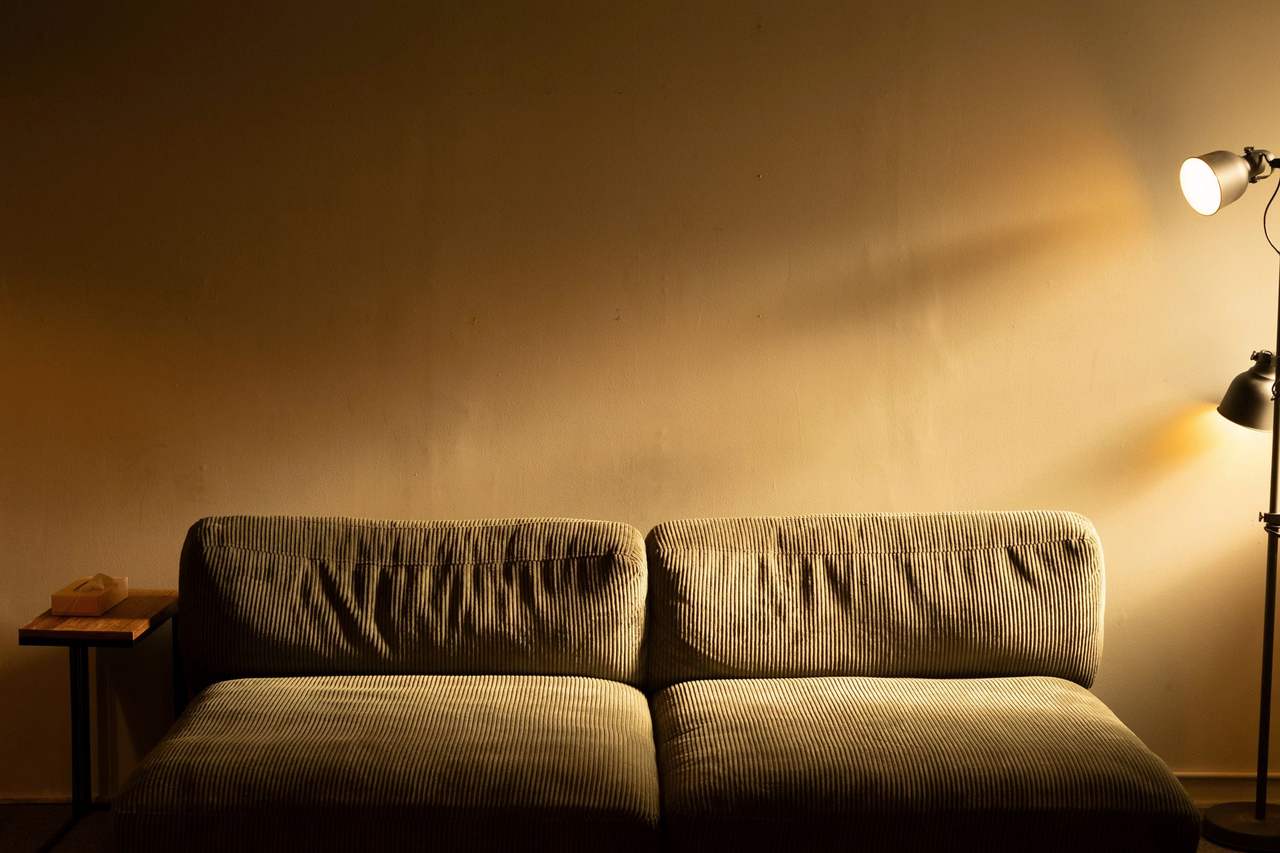 Phòng khách tối với sofa nhung nâu sát bức tường trống, một đèn cây bên phải hắt vệt sáng vàng lên tường