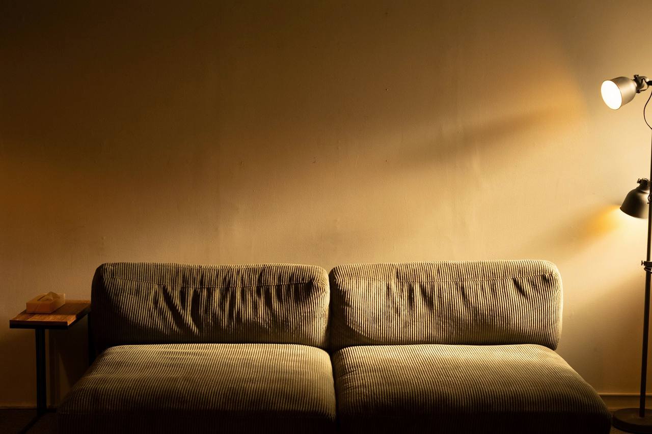 Phòng khách tối với sofa nhung nâu sát bức tường trống, một đèn cây bên phải hắt vệt sáng vàng lên tường