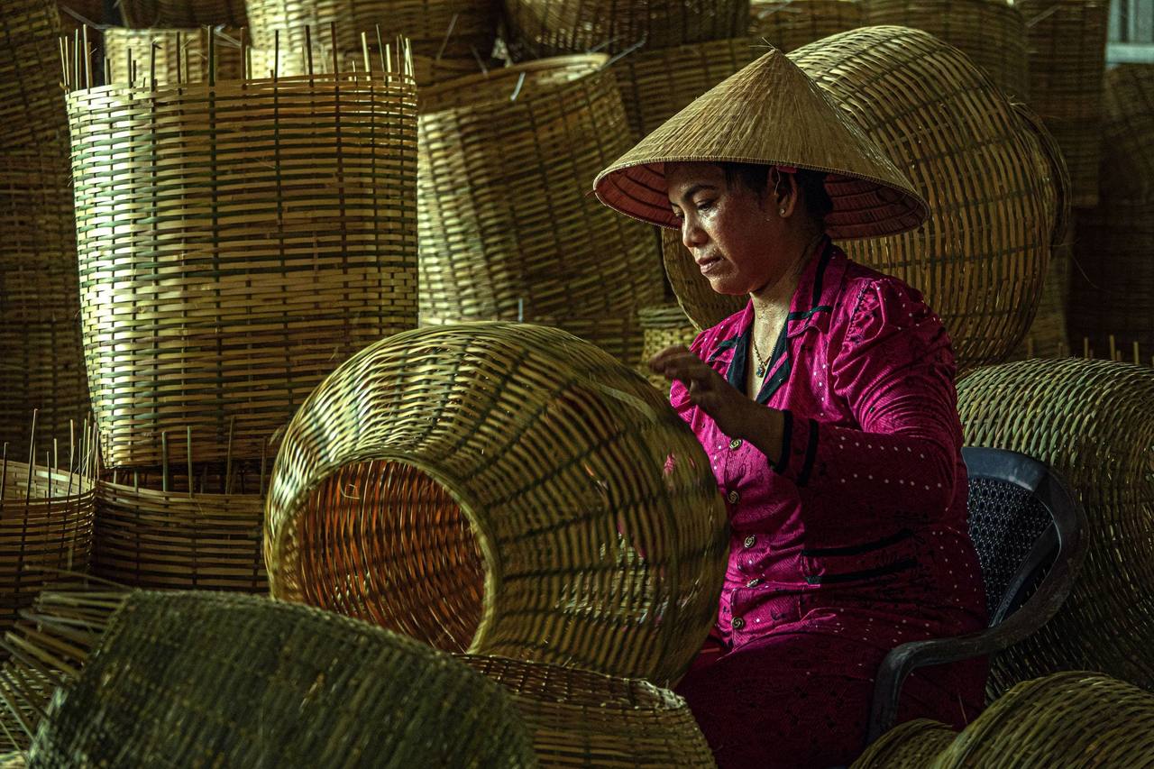 Người phụ nữ Việt Nam đội nón lá, mặc áo hồng, ngồi đan khung tre giữa xưởng chất đầy giỏ đan