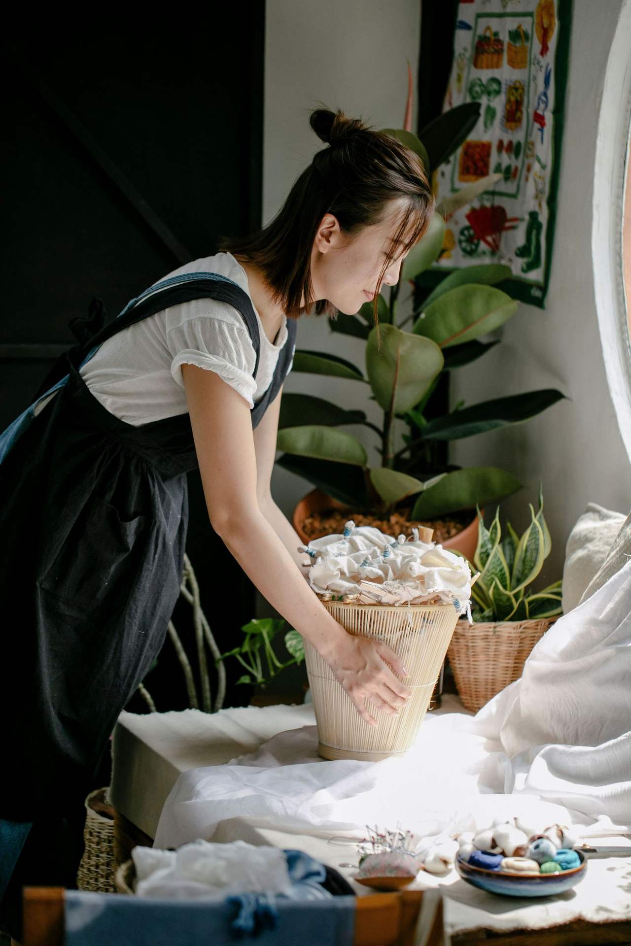 Cô gái trẻ người châu Á đeo tạp dề đen, cúi xuống sắp đồ thủ công trong giỏ đan bên bàn đầy cây xanh
