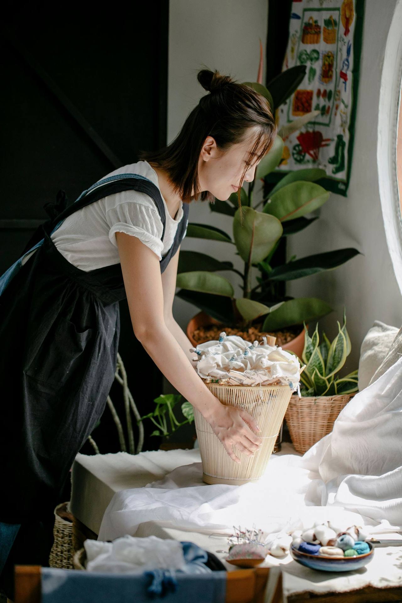 Cô gái trẻ người châu Á đeo tạp dề đen, cúi xuống sắp đồ thủ công trong giỏ đan bên bàn đầy cây xanh