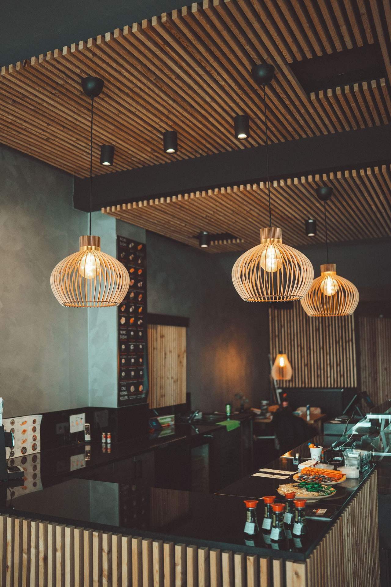 Quầy nhà hàng đá đen dưới trần lam gỗ, hàng đèn thả nan gỗ dáng tròn sáng ấm phía trên